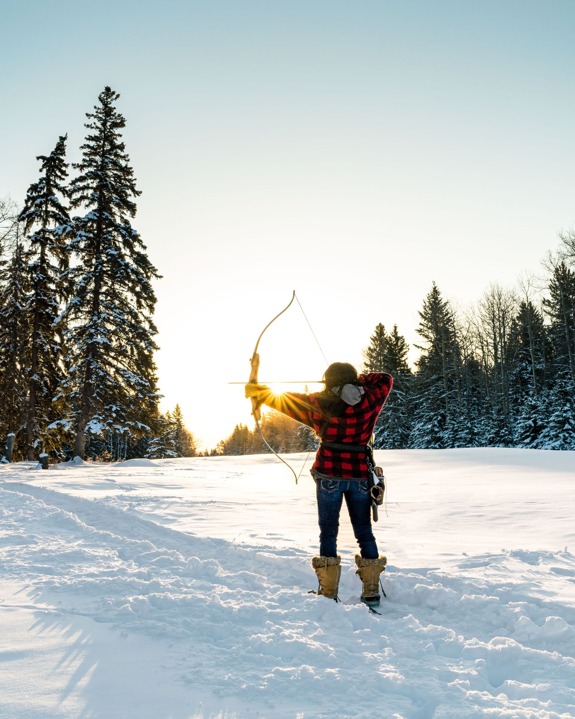 An archer aims a bow into the distance on a winter day in a snowy field, with the sun peeking past her arm.