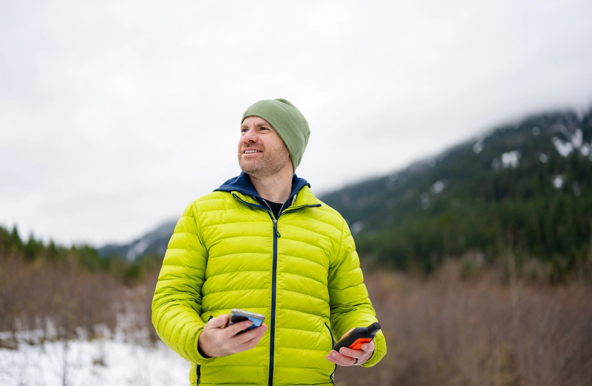 A man outdoors in the winter holding a cell phone in one hand and a satellite communication device in the other.