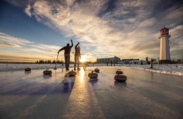 People curling on a frozen lake near a lighthouse in the setting sun.