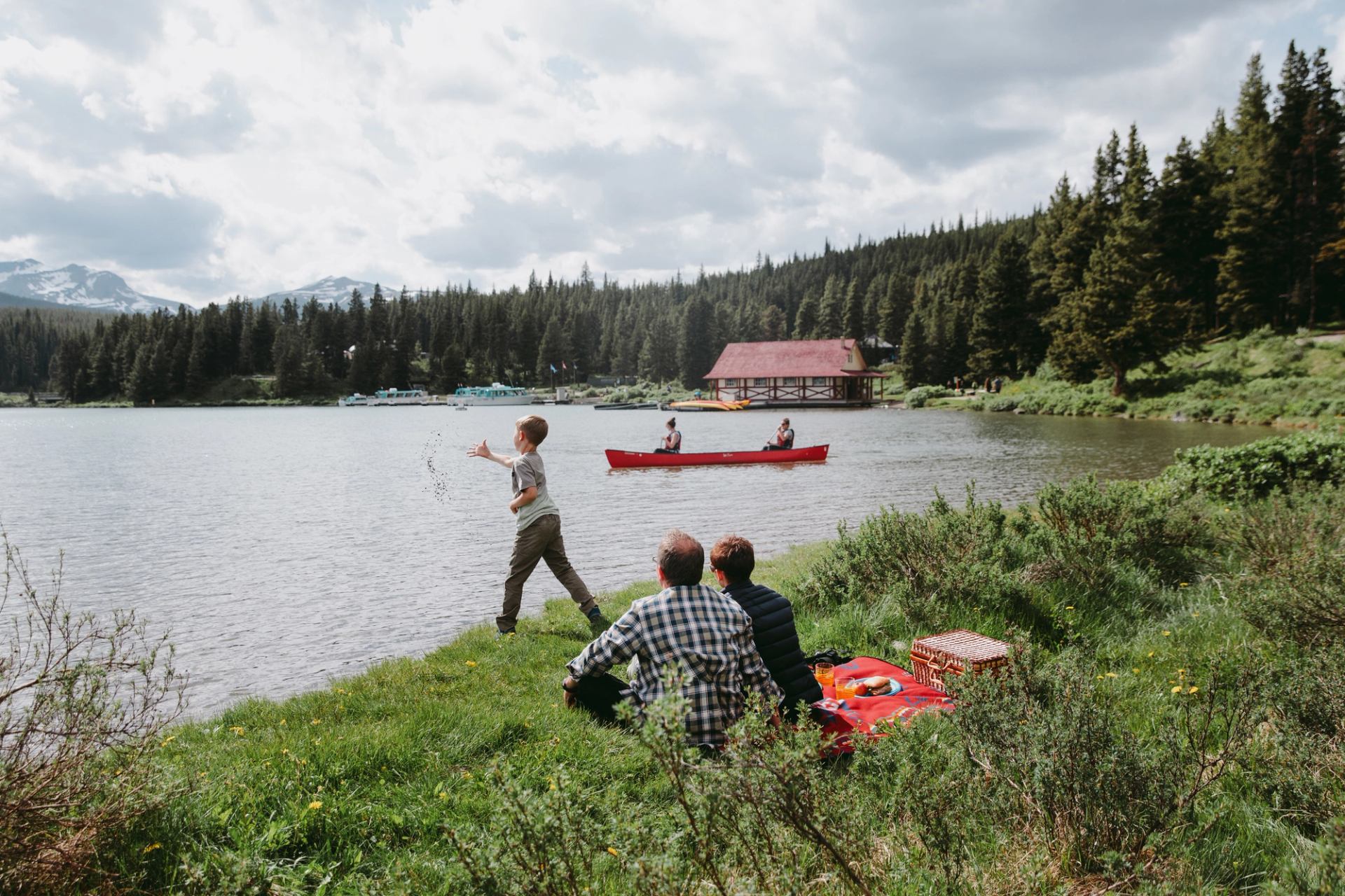 Two parents sitting on a picnic blanket next to a lake watching their son toss rocks into the water.