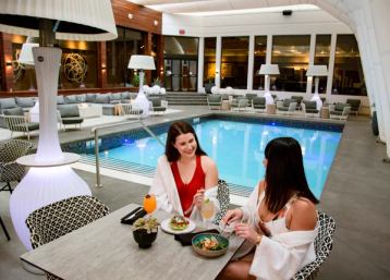 Two women in bathing attire sitting poolside enjoying drinks and food, making for a very trendy atmosphere