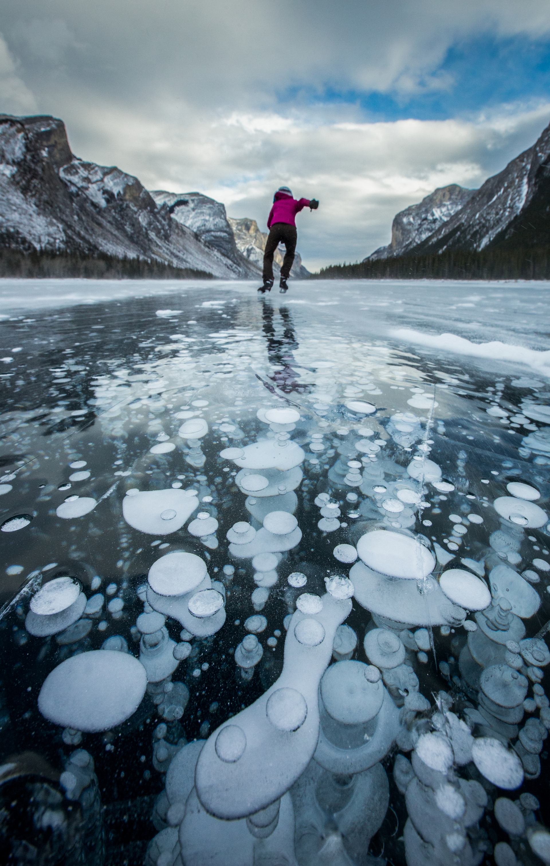 A person skates across Lake Minnewanka and ice bubbles are visible below the surface.