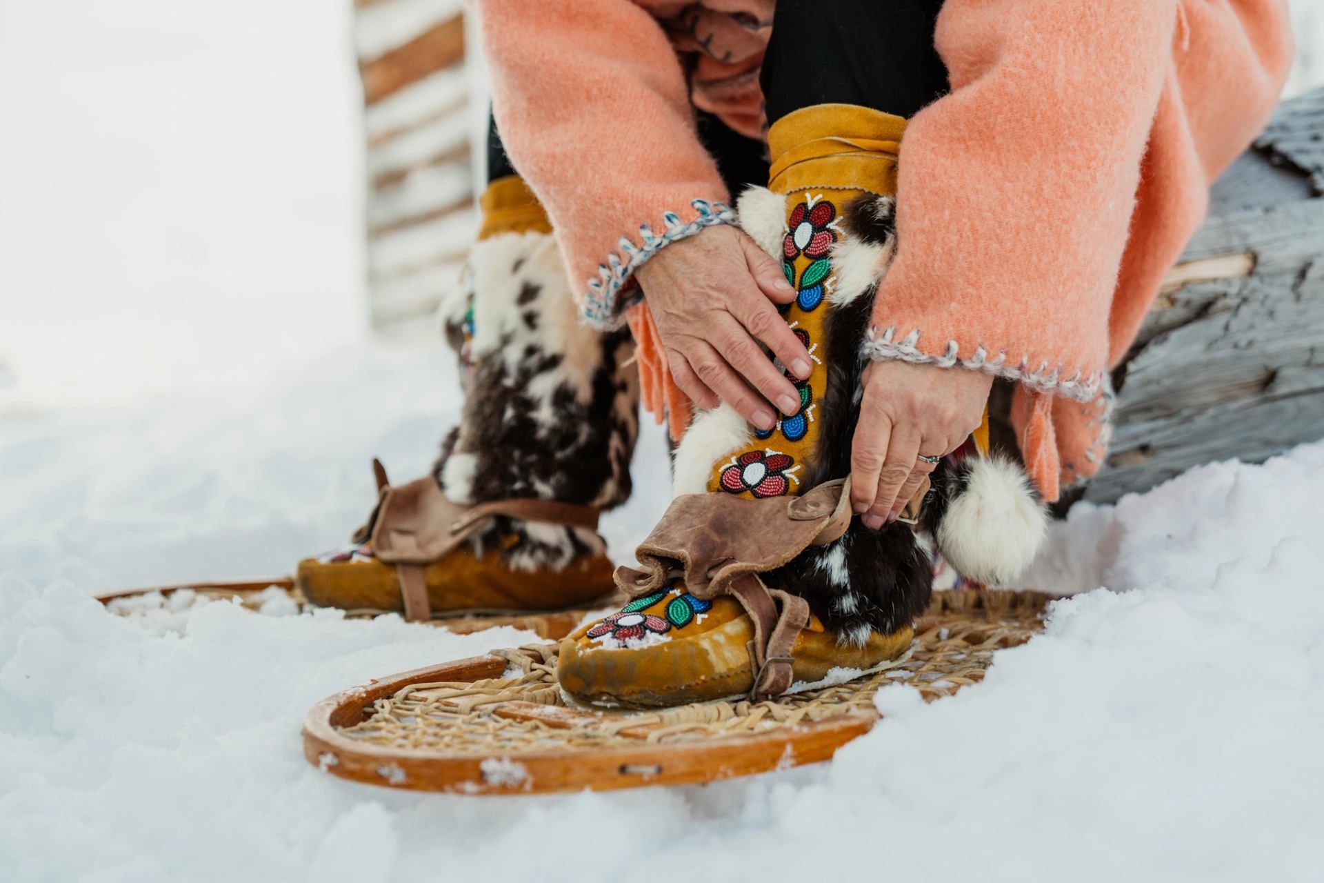 An Indigenous woman in a peach-coloured traditional coat attaches snowshoes to moccasins.