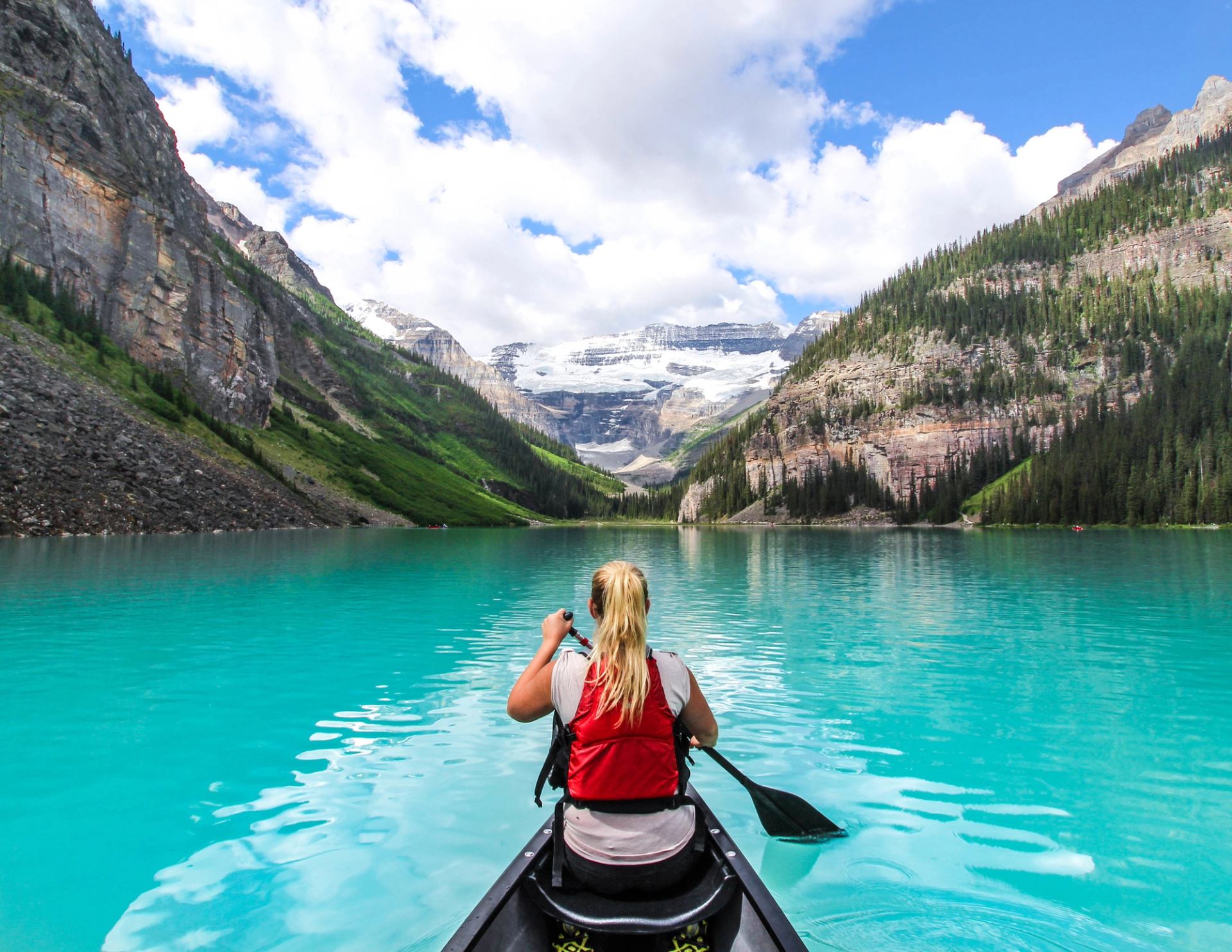 A woman wearing a life jacket paddles a canoe across the turquoise water of Lake Louise with two mountains in the distance meeting in a valley.
