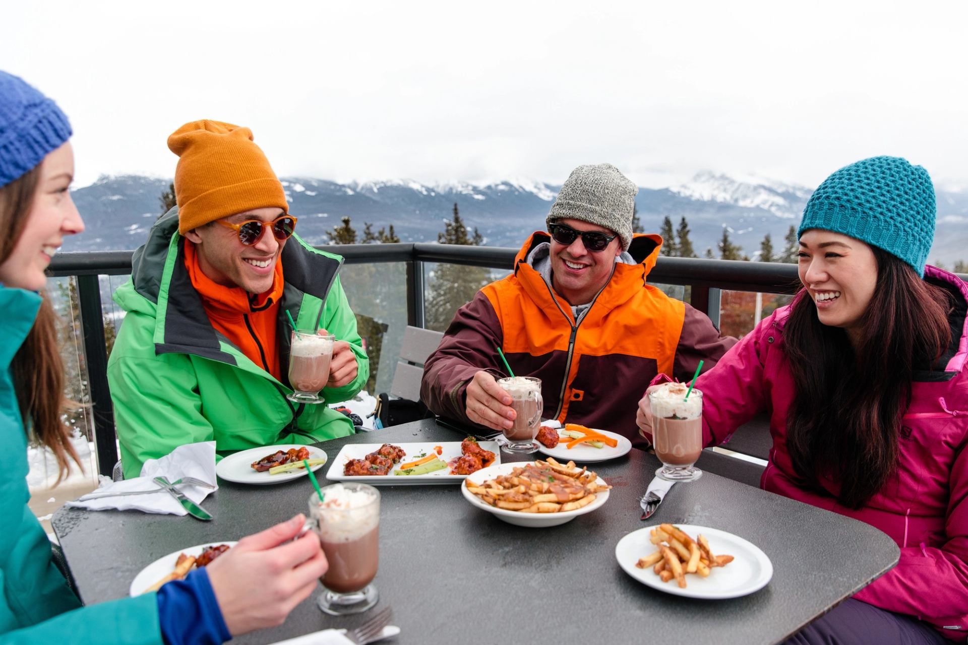A group of four skiers sit outside on a slopeside patio, enjoying food and drinks, after a day of skiing Marmot Basin in Jasper.
