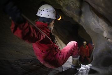 A women propelling herself down into a dark cave lit by her headlamp with people watching from below.