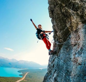 Rock climber waving off the side of a mountain headwall with a lake view in the background