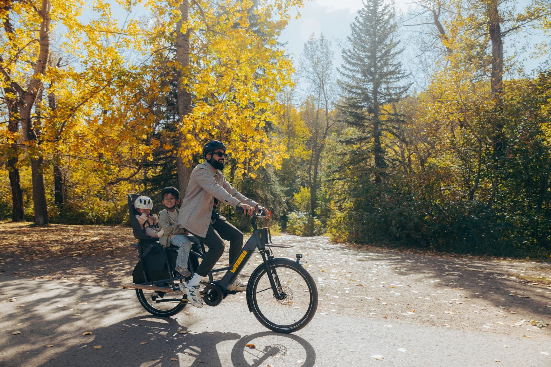A smiling dad rides an e-cargo bike past trees with golden leaves along a paved pathway