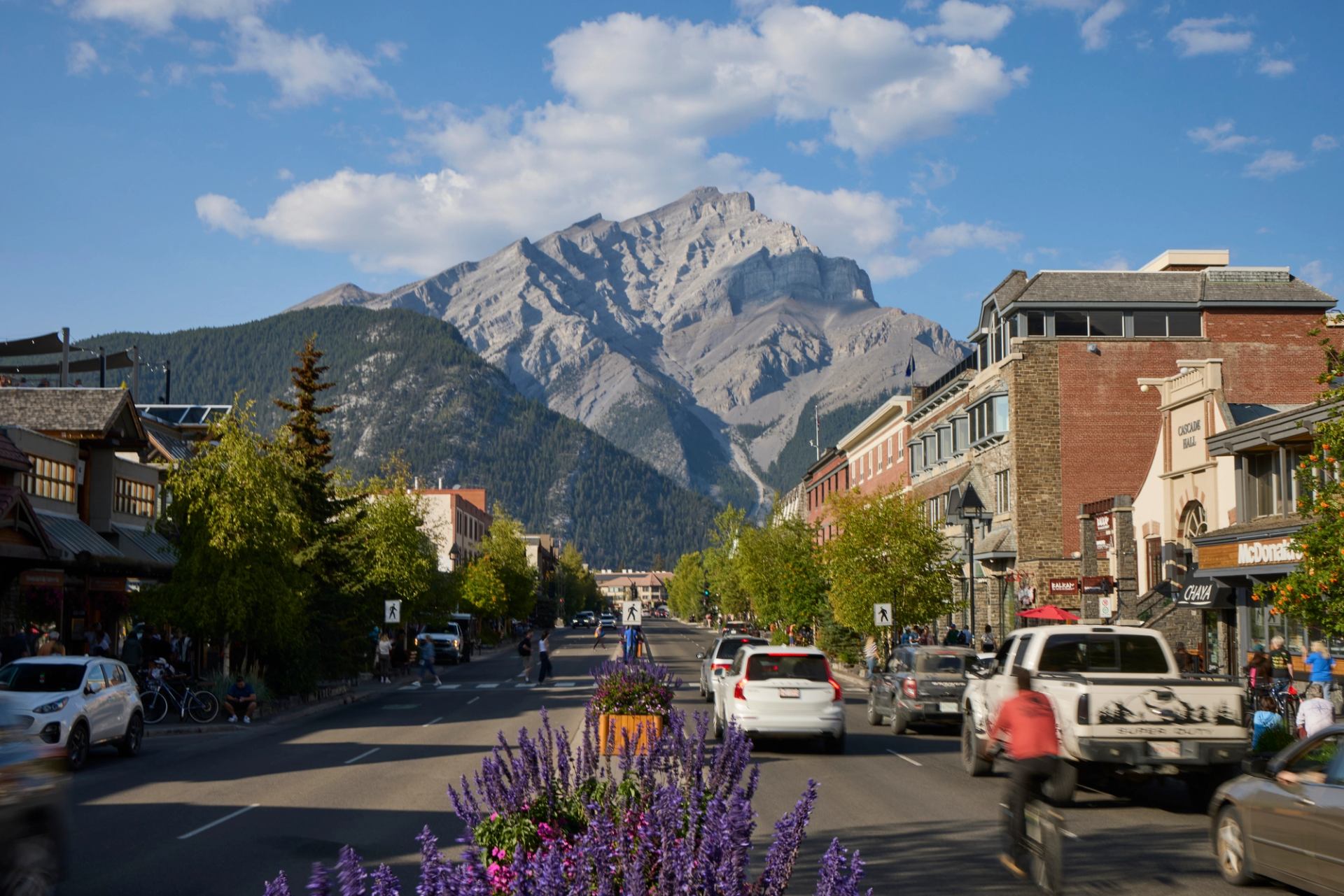 A view down Banff Ave.  with Cascade Mountain rising in the distance, cars and cyclists on the road, and shops lining the street.