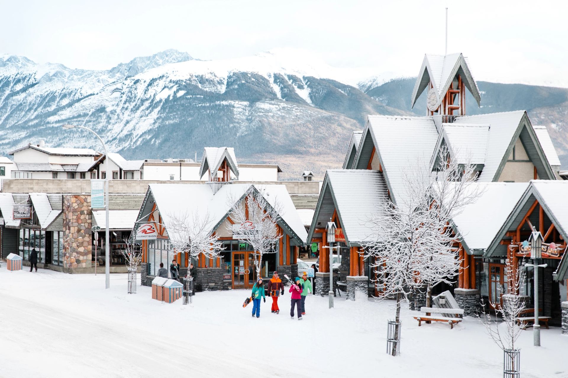 Skiers and snowboarders walking in downtown Jasper, with buildings and shops in the background.