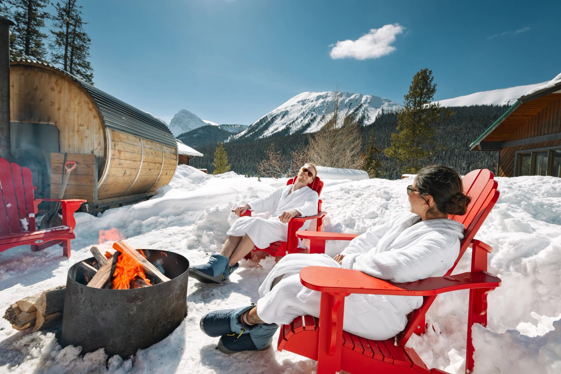Couple relaxing in red Adirondack chairs by an outdoor fire pit, wearing robes and sunglasses, enjoying sunshine and mountain views.