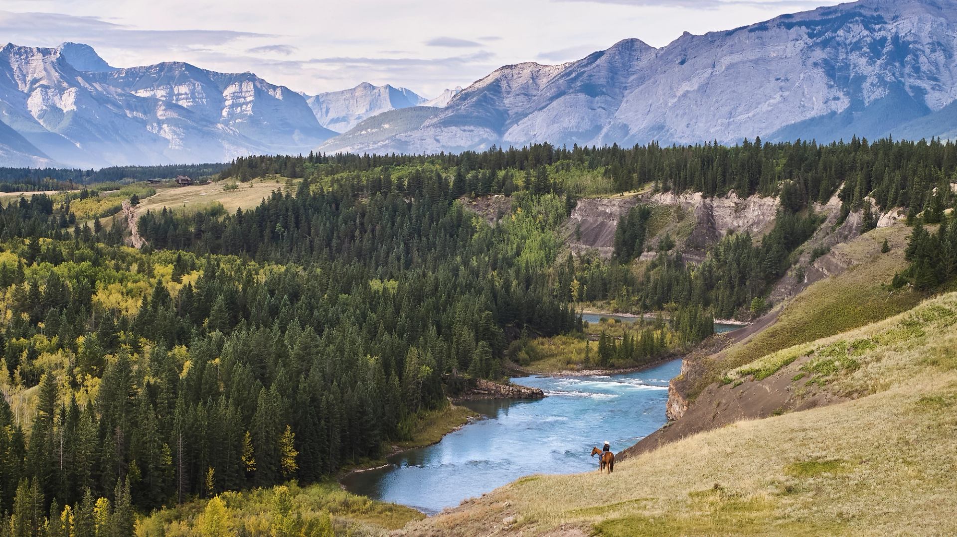 Alberta’s Foothills featuring distant Rocky Mountains, rolling foothills and a horseback rider near a river.