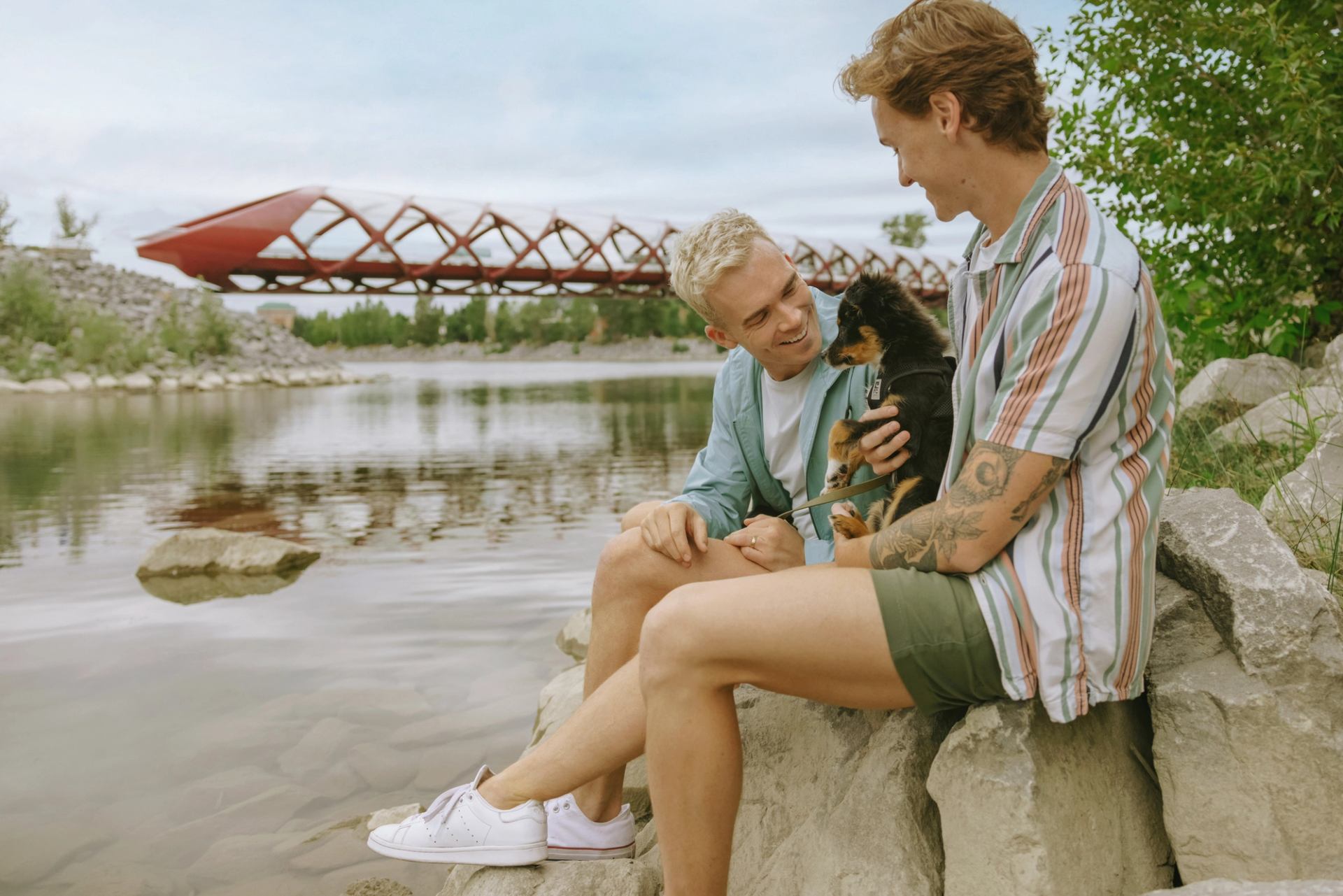 Two people with a puppy sit along the river with a bridge in the background.