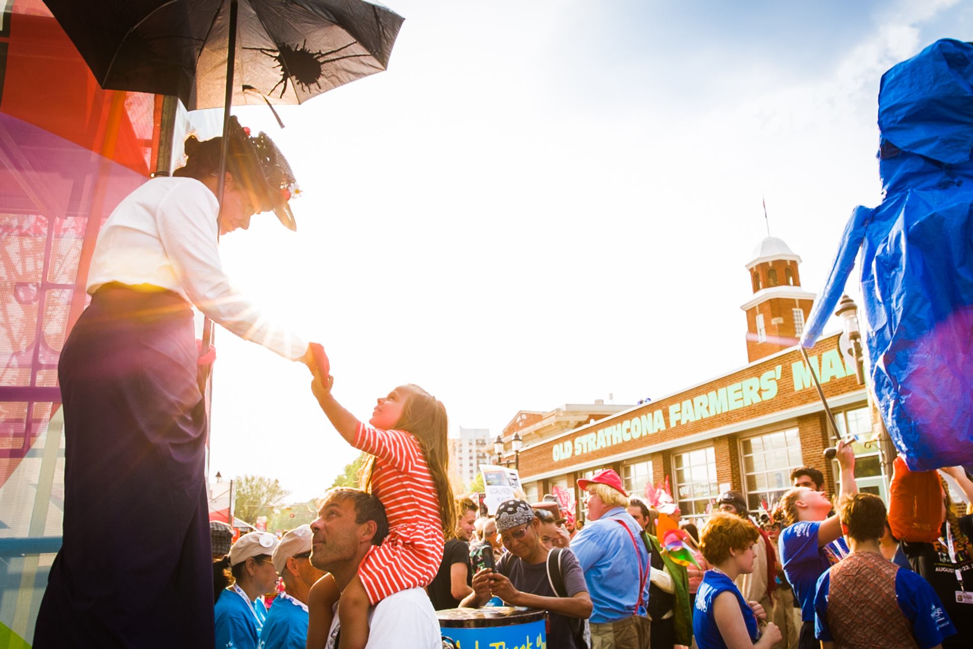 A young girl sitting on her father's shoulders interacts with a performer on stilts.