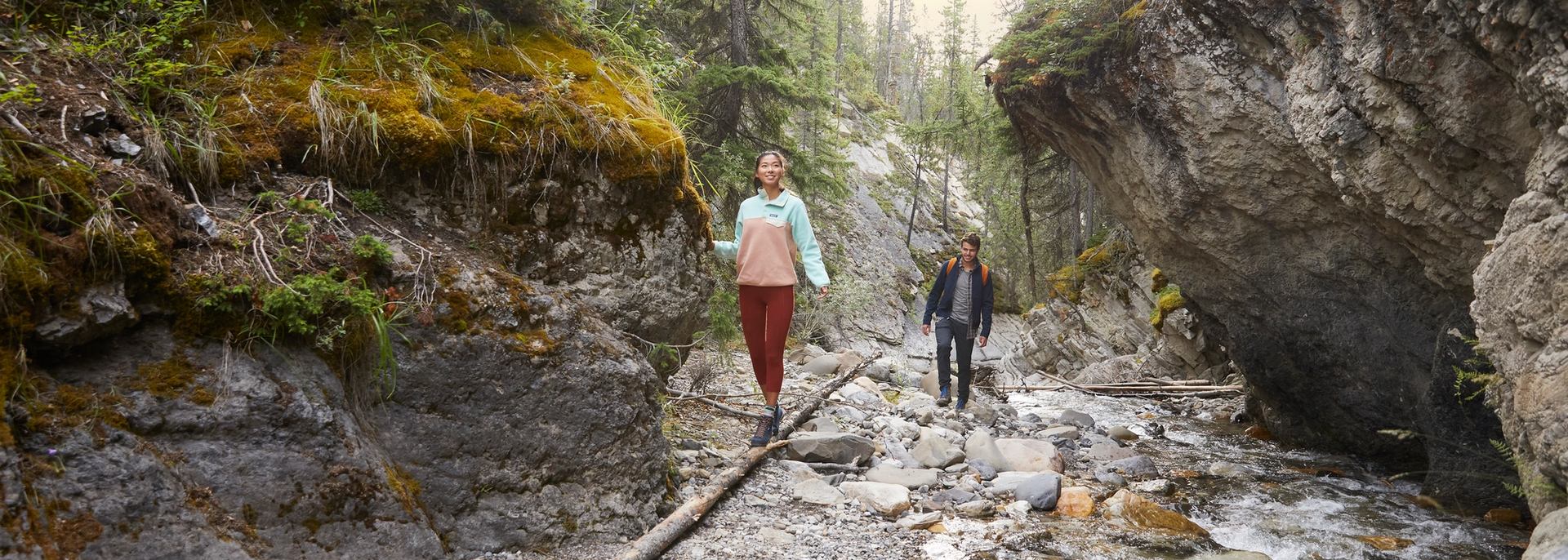 A woman balancing on a fallen down tree trunk with a man behind her standing on rocks as they are on a hike together admiring Whitegoat Falls in Nordegg.