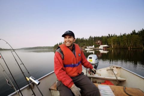 Owner and fishing guide of Andrew Lake Lodge driving a boat on a lake with calm waters and the lodge in the background.