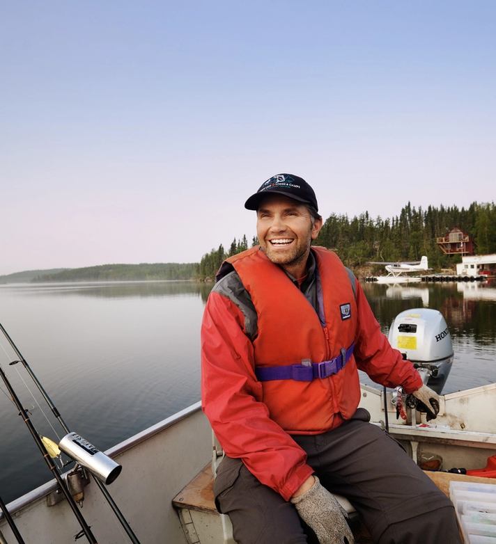 Owner and fishing guide of Andrew Lake Lodge driving a boat on a lake with calm waters and the lodge in the background.
