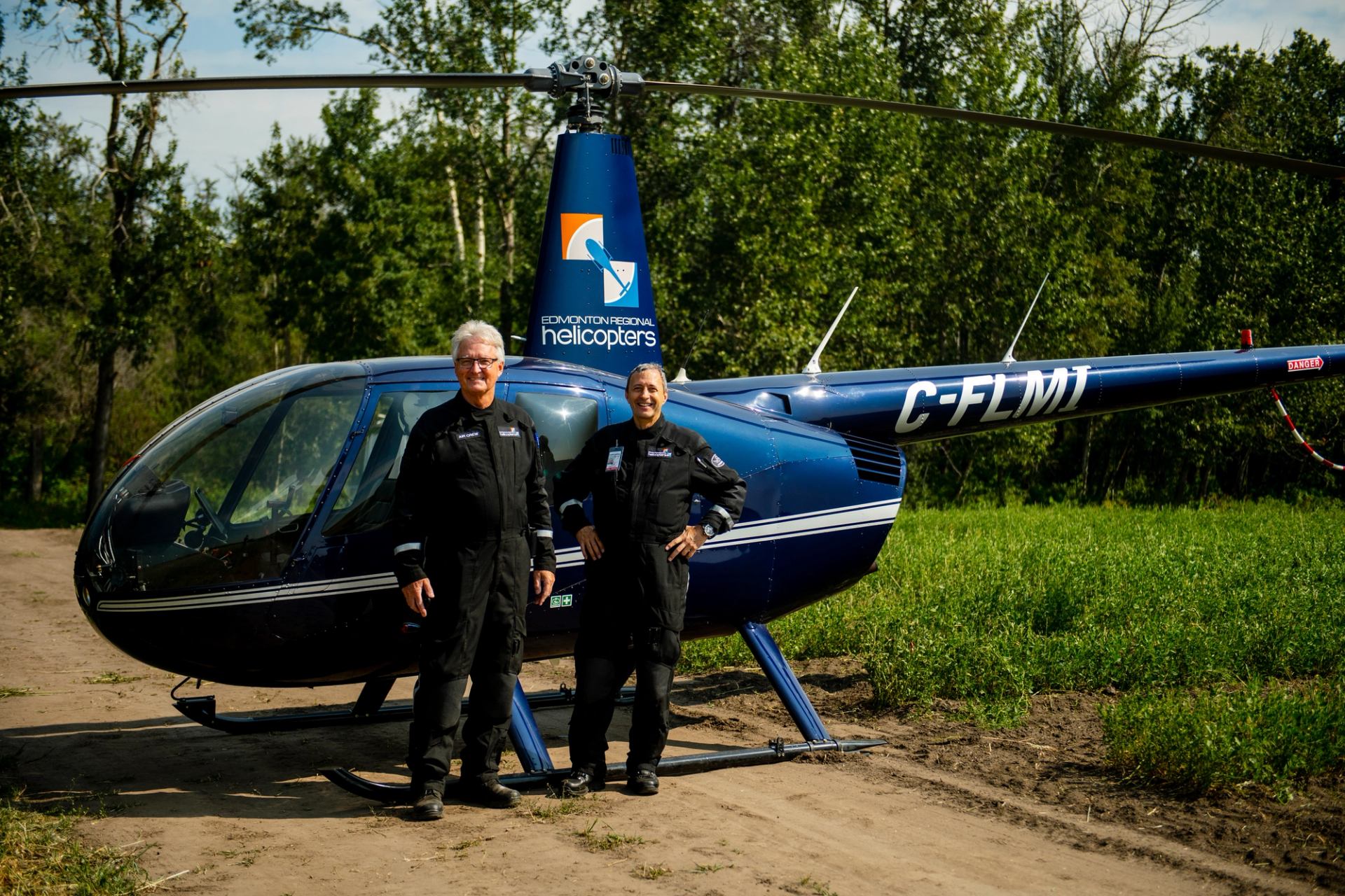 Two men in pilot jumpsuits standing in front of a helicopter on a remote dirt road