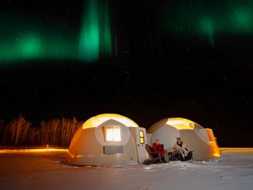 Couple sitting in front of glamping domes at Metis Crossing under the aurora borealis, northern lights night sky