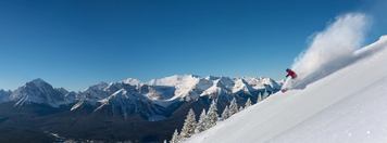 A skier in a yellow jacket carves through powder on a mountain top with the Canadian Rockies in the distance.