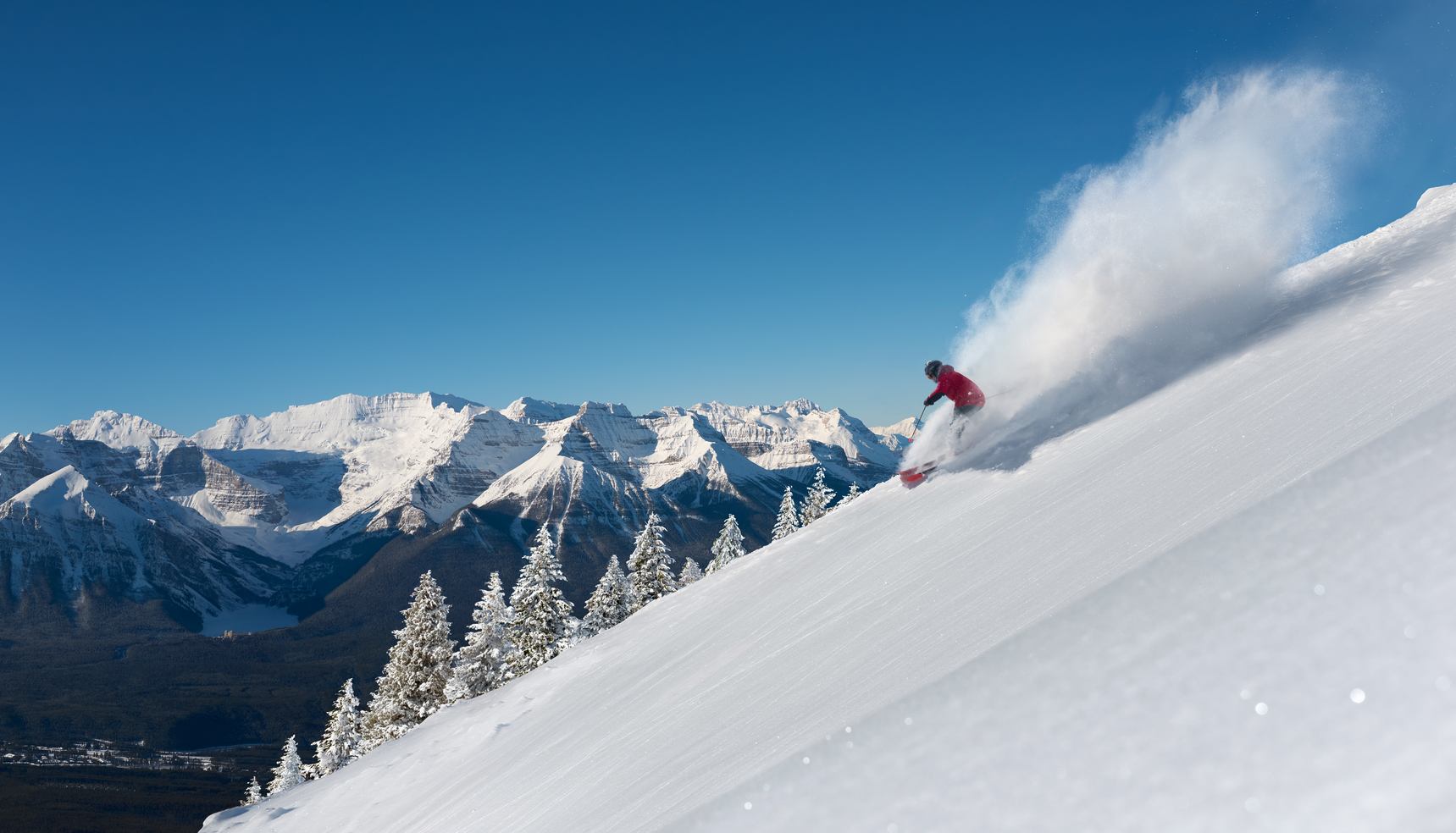 A person skiing down a mountain at Lake Louise Ski Resort, carving deep powder under blue skies.