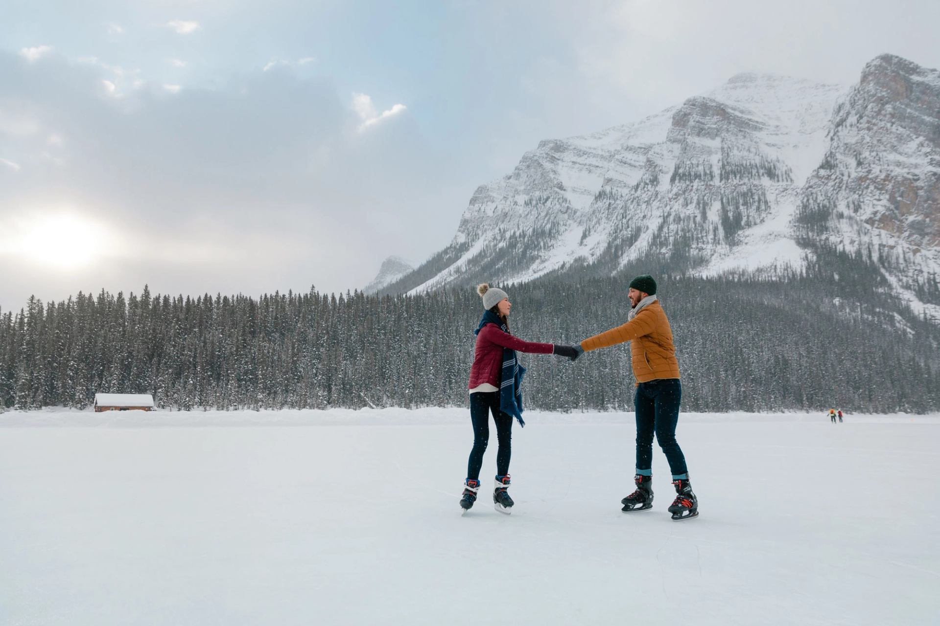 A couple holding hands while ice skating on Lake Louise, mountains and trees in the background, in Banff National Park.