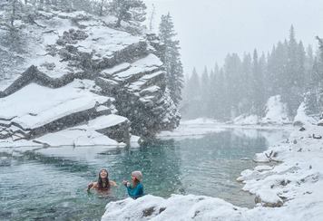 Two women cold plunging in a mountain lake on a snowy day at Canoe Meadows in Alberta's mountains.