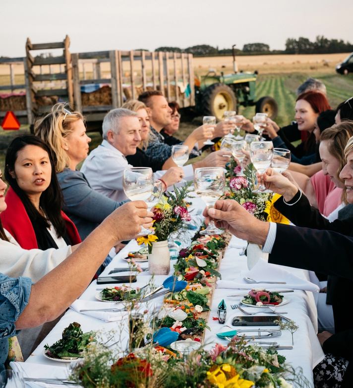 Large group clinking glasses at an outdoor dinner table on a farm.