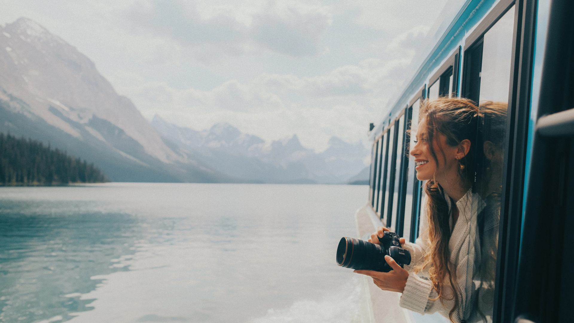 A woman leans out the window of the Maligne Lake Cruise boat with her camera to look at mountains surrounding the lake.