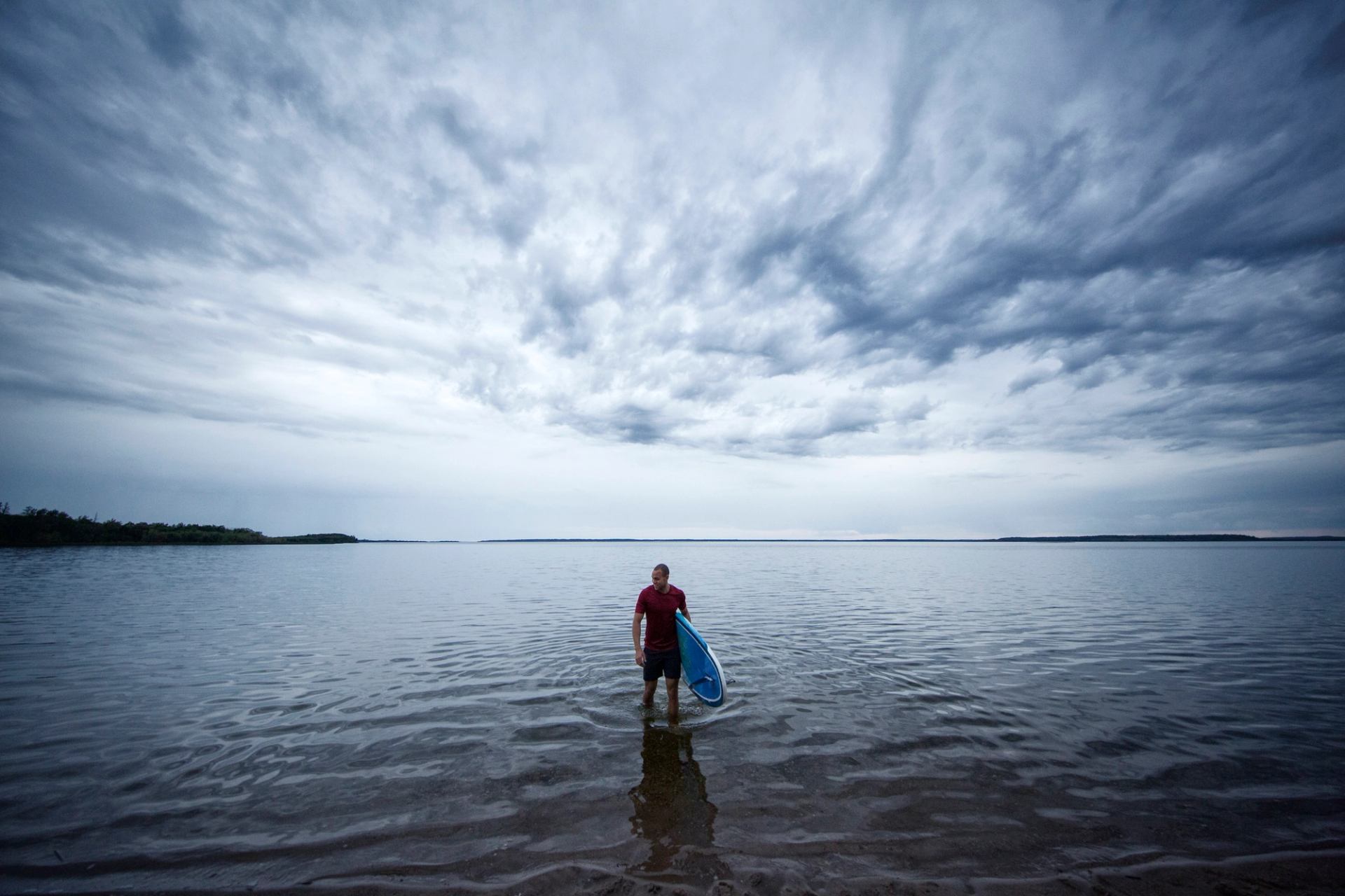 A paddleboarder walks to shore carrying his board under moody skies.