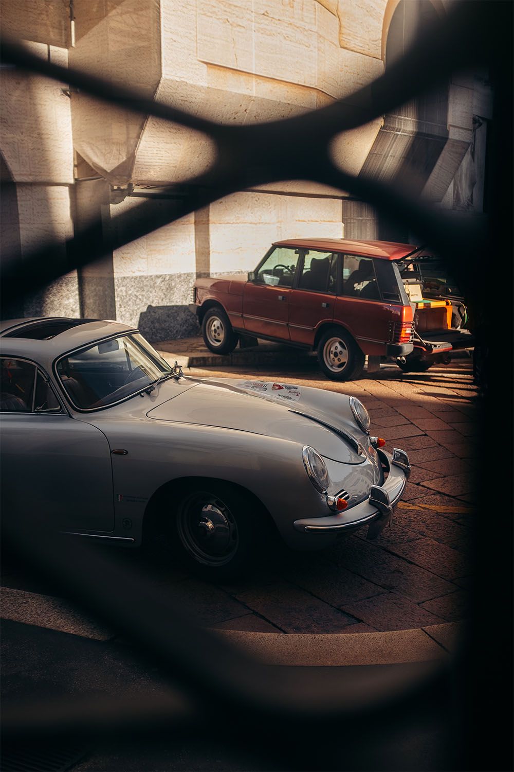 Porsche 356 argento in primo piano e Range Rover classico rosso sullo sfondo, fotografati attraverso una grata in un contesto urbano.