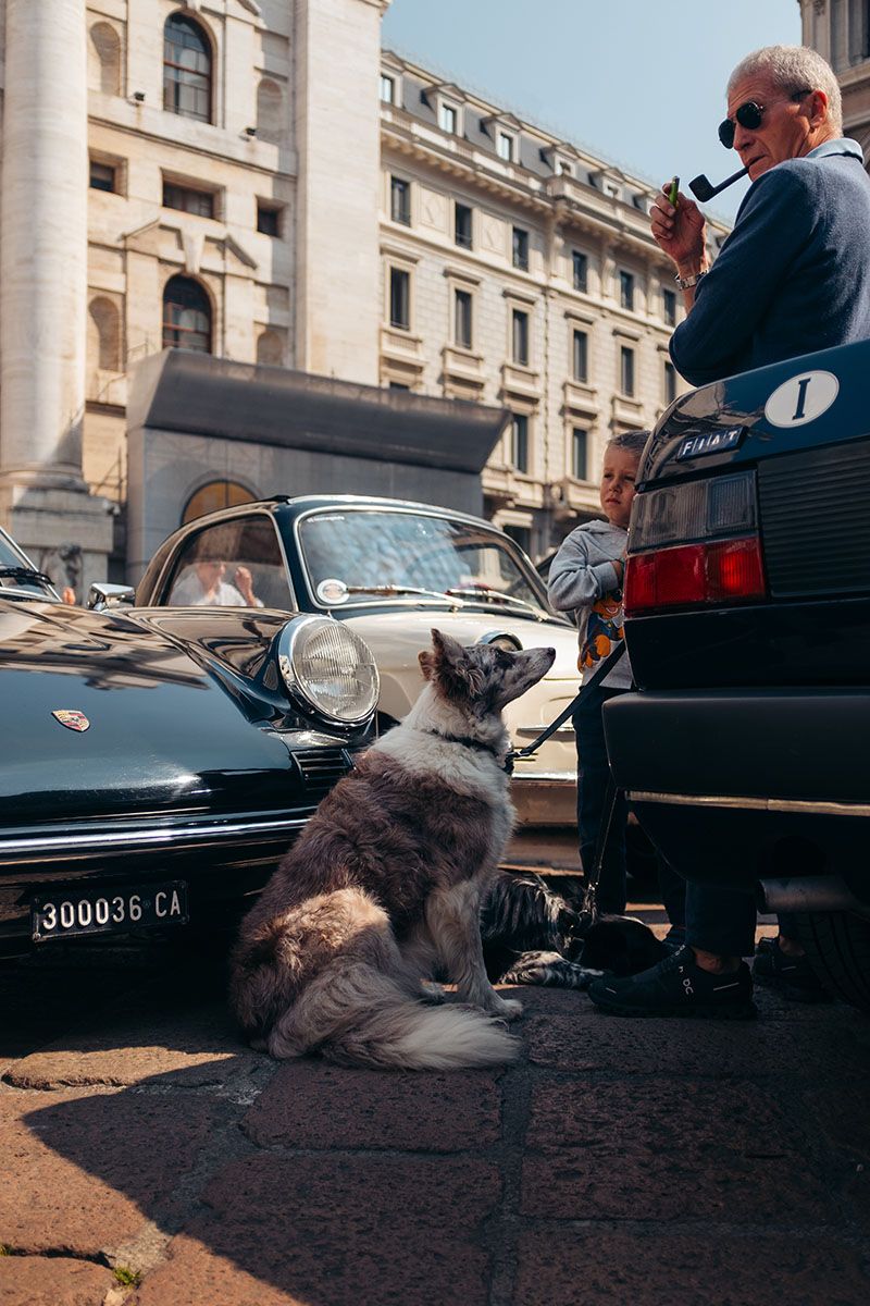 Uomo con pipa, bambino e cane accanto a un’auto classica in Piazza Affari durante un evento automobilistico.