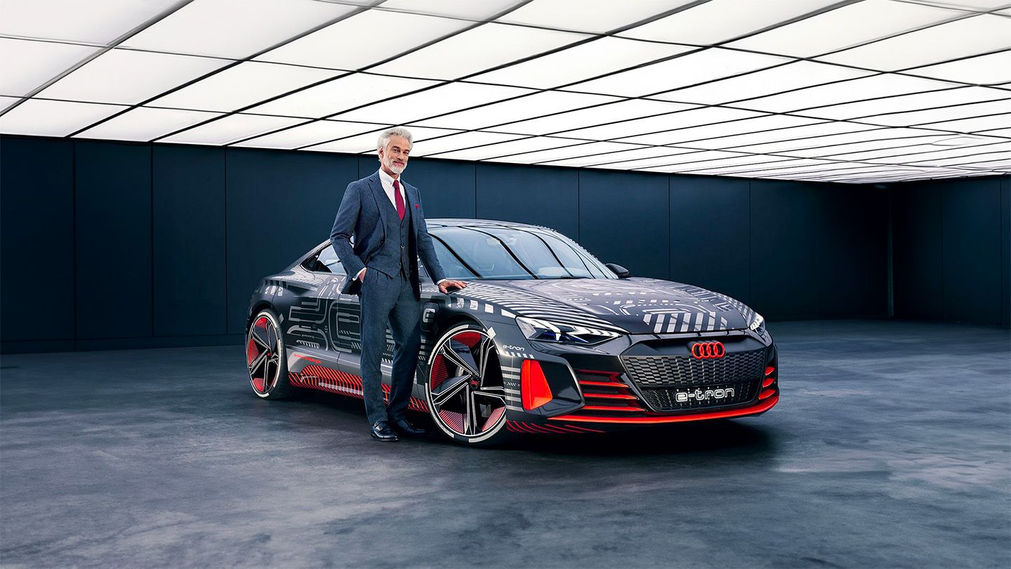 Elegant businessman in a formal suit poses next to a modern Audi e-tron GT electric car in a dimly lit indoor display, representing a festive advertisement that combines professional style and automotive innovation.