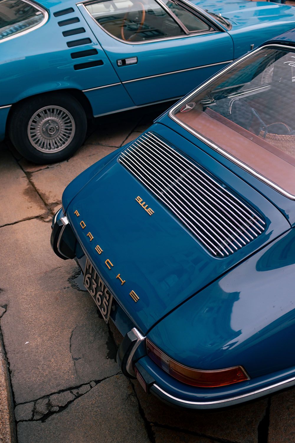 Vista dall’alto del posteriore di una Porsche 911 blu con scritta dorata “911e”, accanto a una coupé classica azzurra.