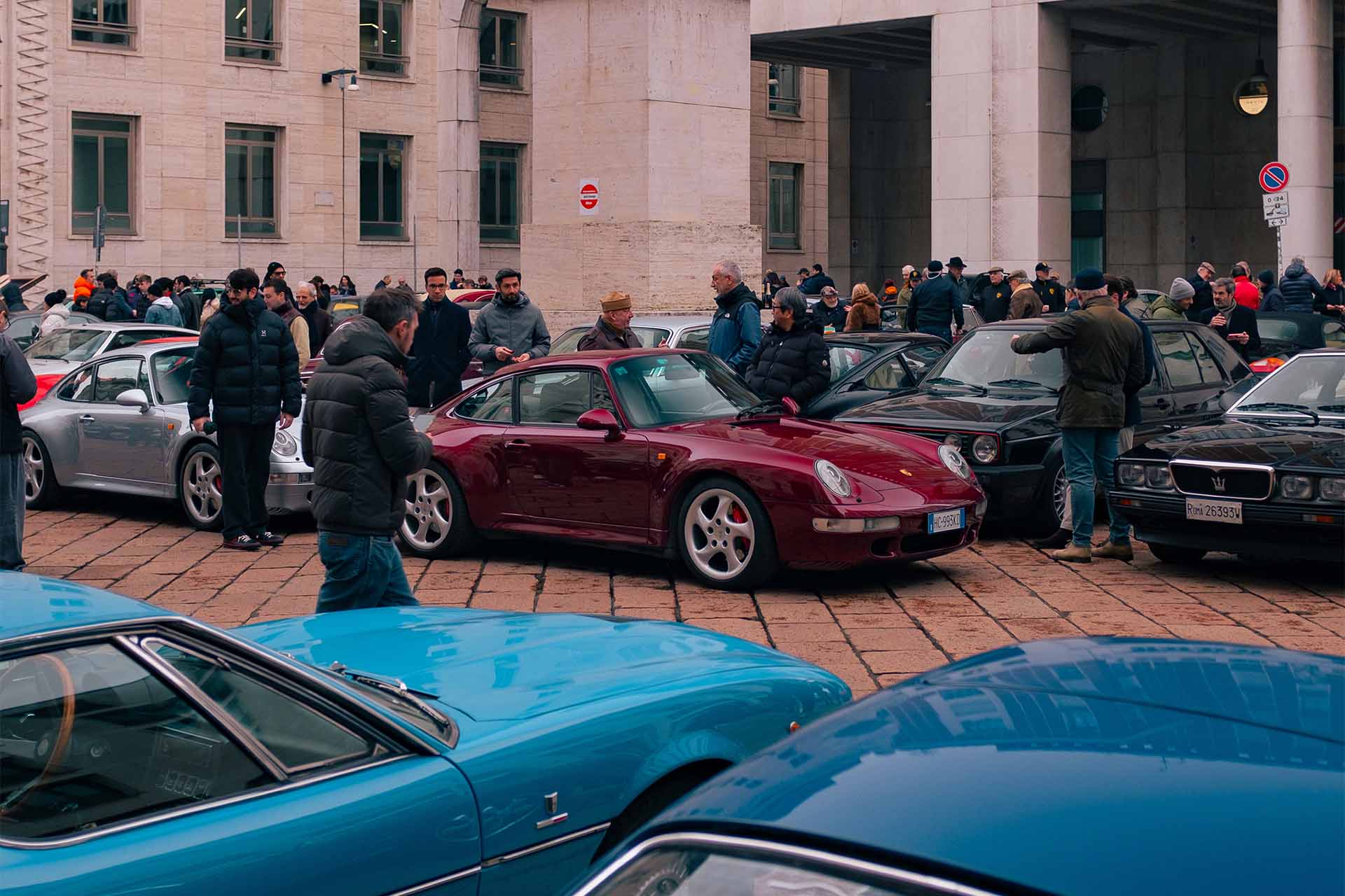 Auto sportive e classiche parcheggiate in Piazza Affari a Milano durante un raduno, con una Porsche 911 bordeaux al centro e persone intorno alle vetture.