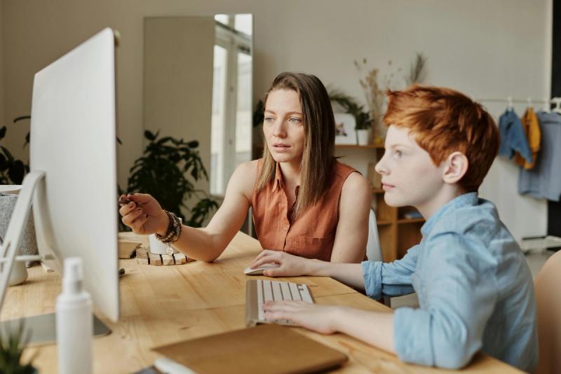 A parent is sitting with a young boy at a desk and explaining something they are both looking at on a laptop screen.
