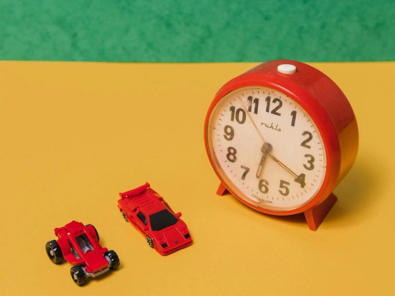 Two red toy racing cars on a yellow table beside a red analogue alarm clock