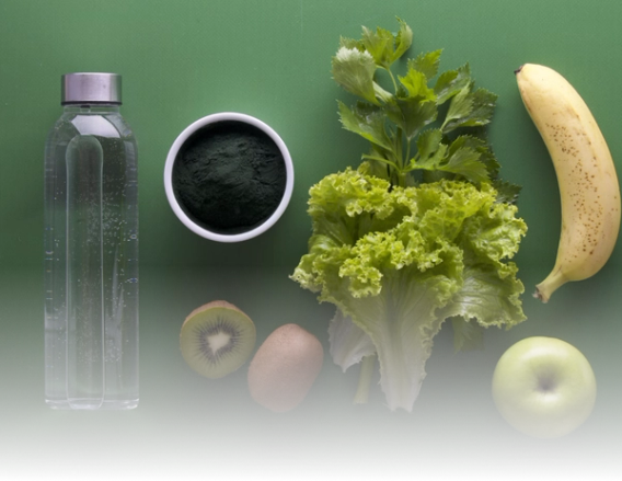 Water bottle, greens powder, kiwi, lettuce, celery, banana, and apple arranged on a green surface