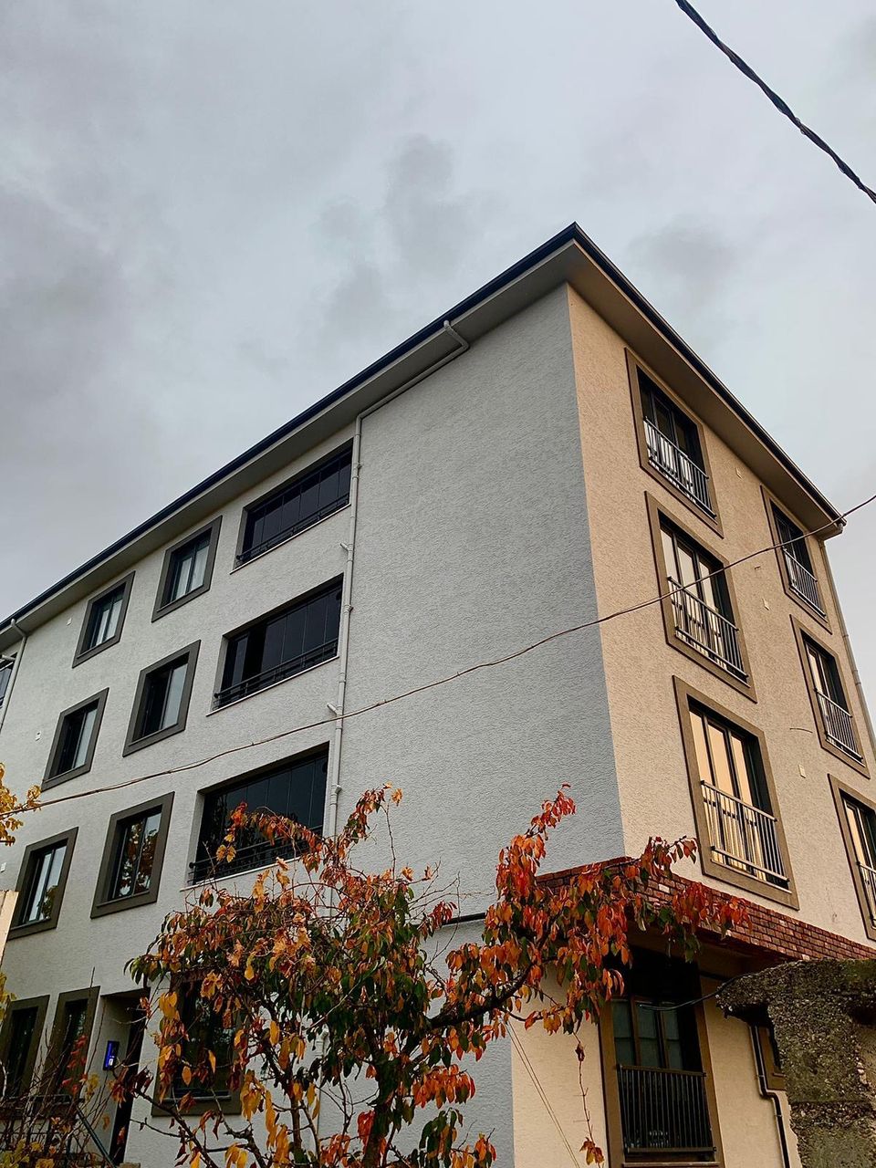 Binanın dış cephesinin gerçek fotoğrafı; kat pencereleri, balkonlar ve ön planda sonbahar yaprakları görülüyor
