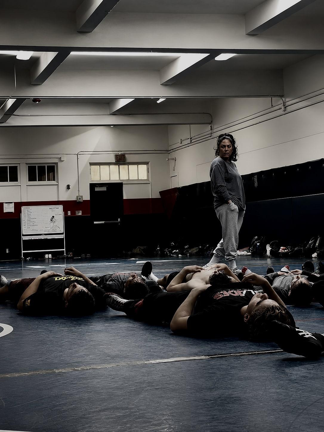 A coach standing over a row of wrestlers lying on the gym mat during a guided mental training session.