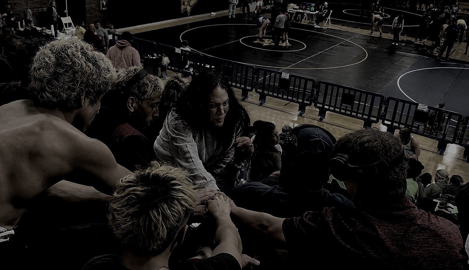 A high school wrestling team huddled close in the stands above the mat, hands stacked together, their coach leaning in mid-sentence just before they compete.