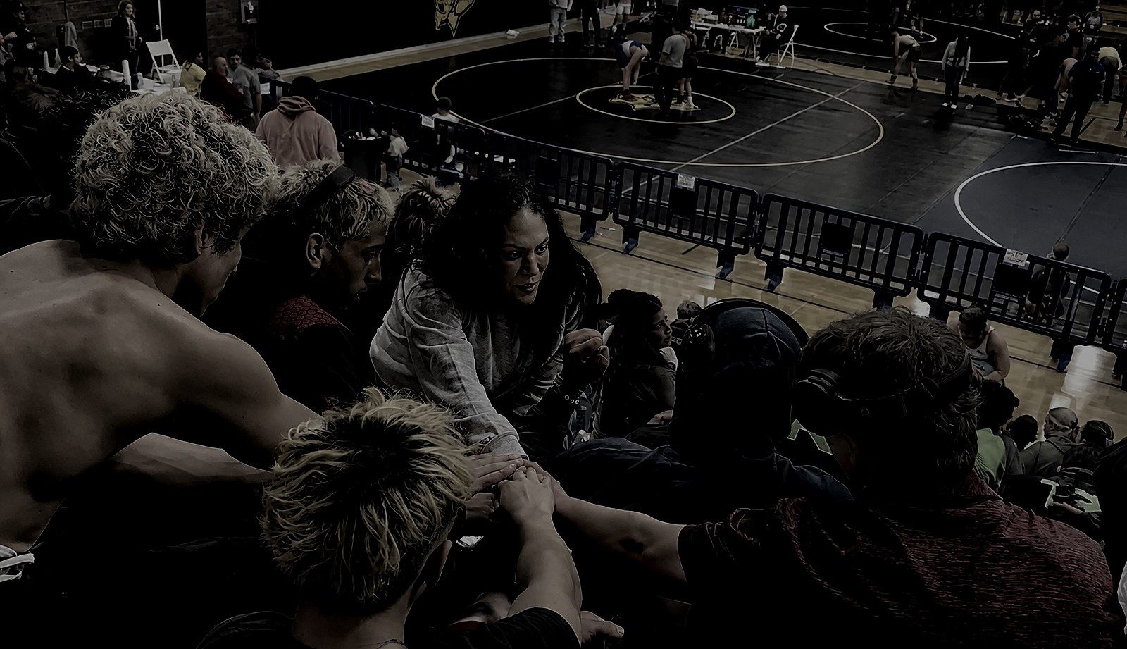 A high school wrestling team huddled close in the stands above the mat, hands stacked together, their coach leaning in mid-sentence just before they compete.