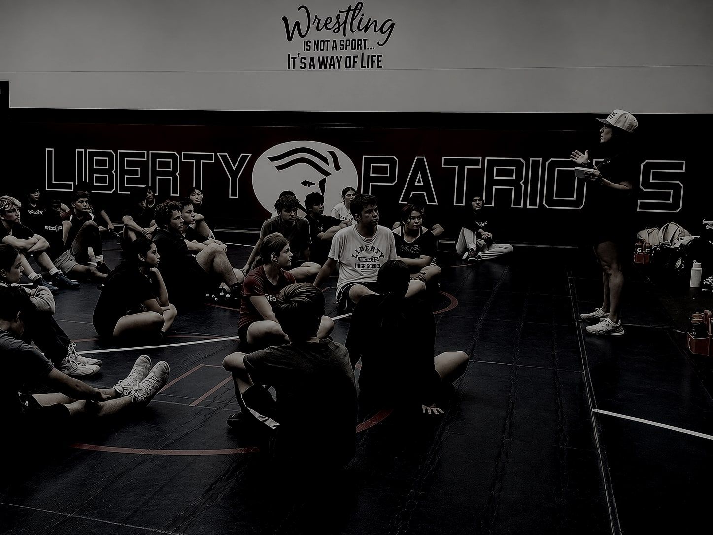 Athletes seated in a circle on a wrestling mat during a team session, under a wall reading "Wrestling is not a sport, it's a way of life."