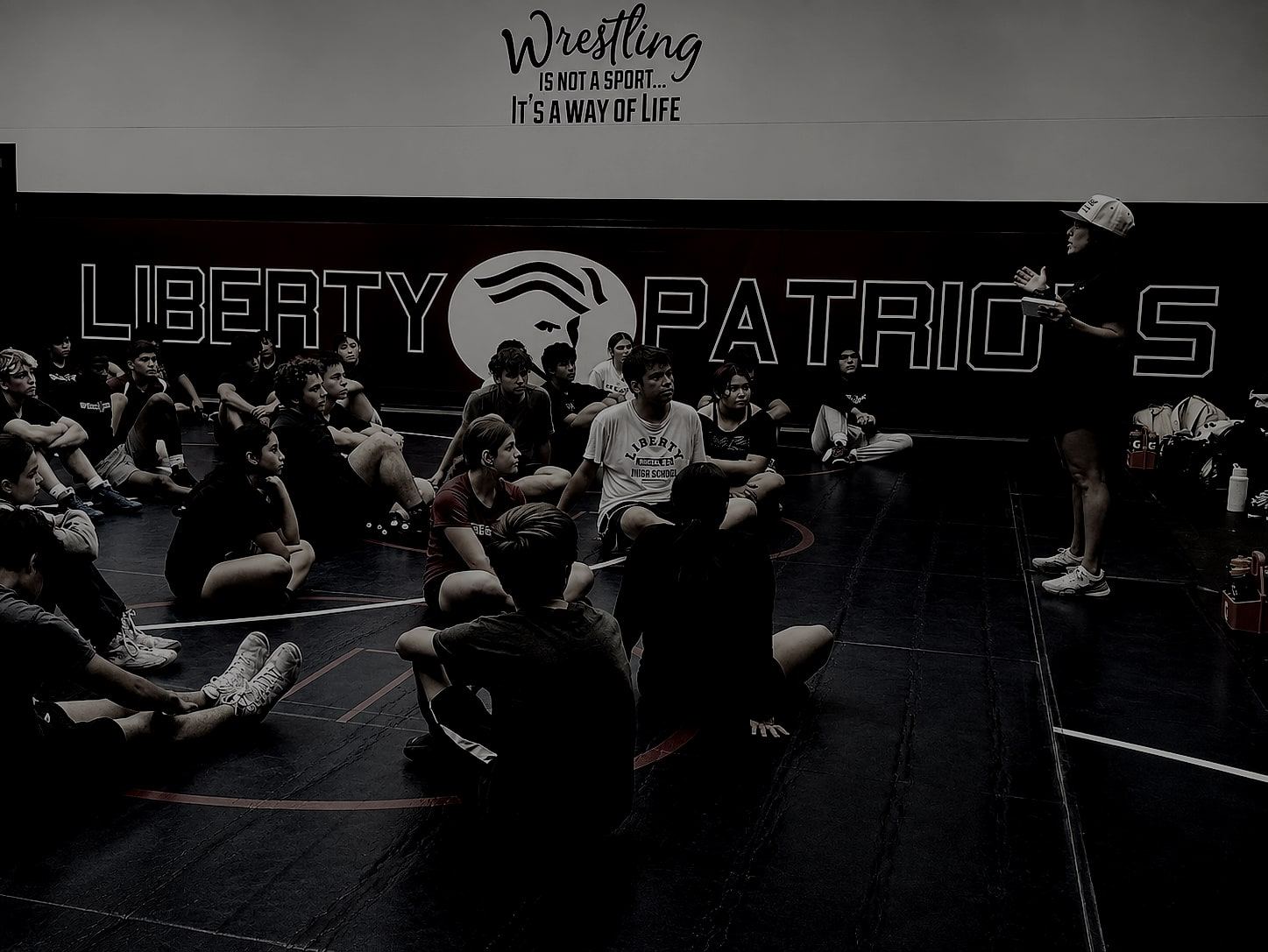 Athletes seated in a circle on a wrestling mat during a team session, under a wall reading "Wrestling is not a sport, it's a way of life."