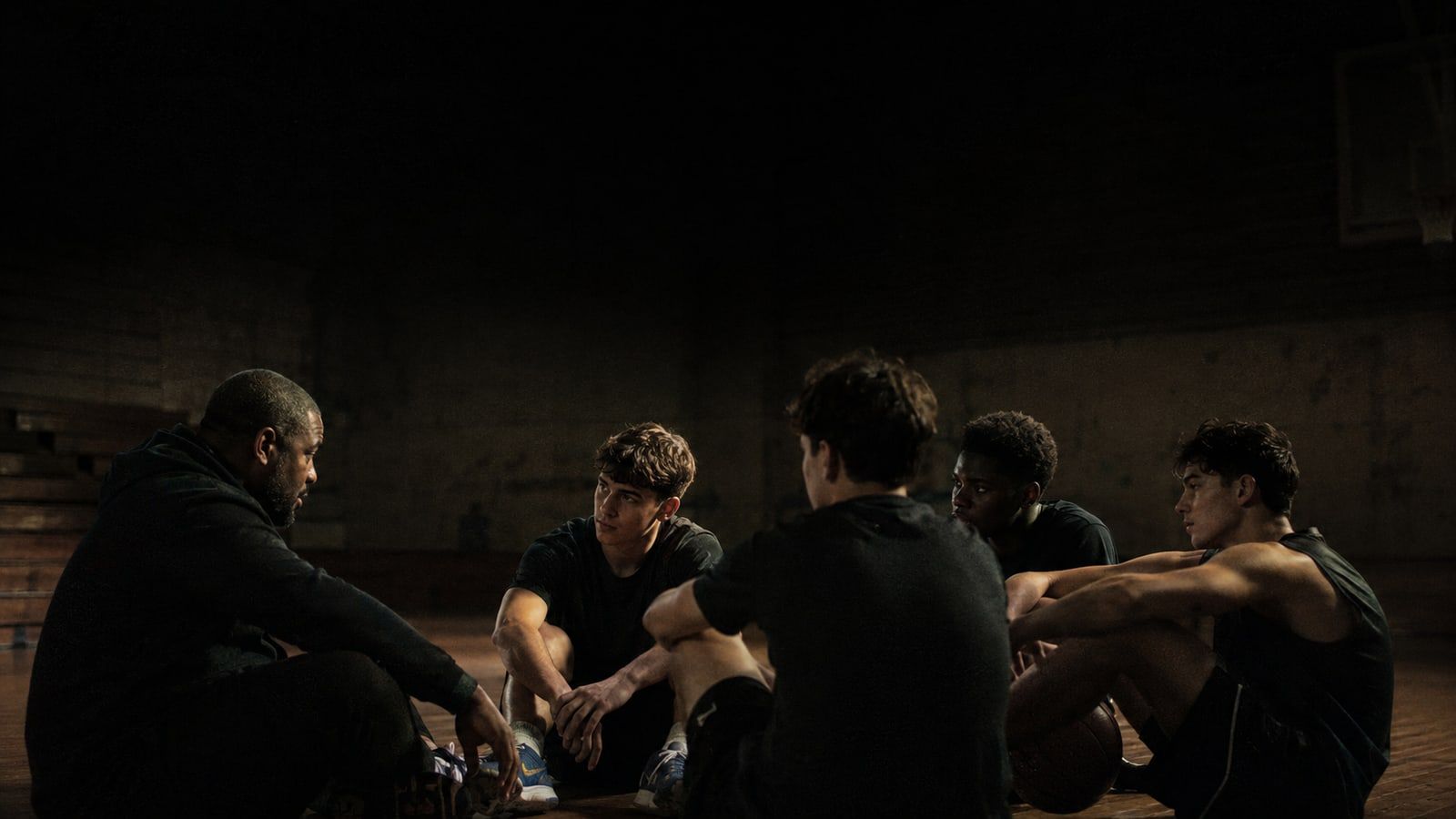 A coach sitting on the gym floor at eye level with five teenage athletes, mid-conversation, in low light.