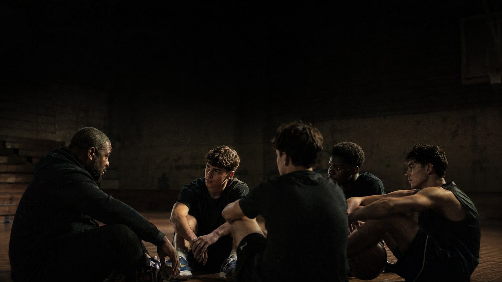 A coach sitting on the gym floor at eye level with five teenage athletes, mid-conversation, in low light.