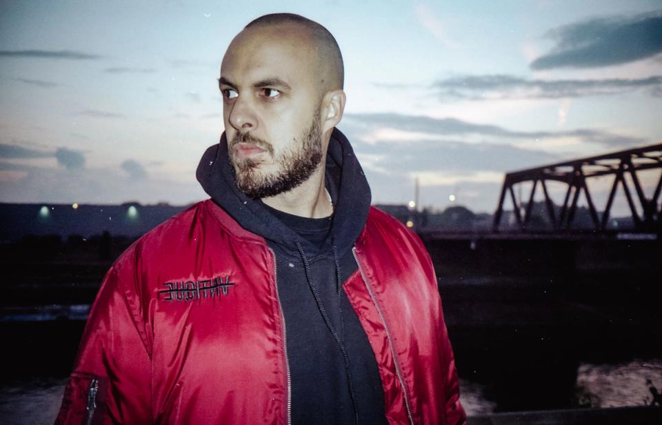 Producer Alexis Troy trägt eine rote Bomberjacke vor einer Brücke