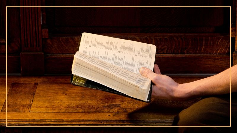 An outstretched hand holding an open Bible over a wood tabletop.