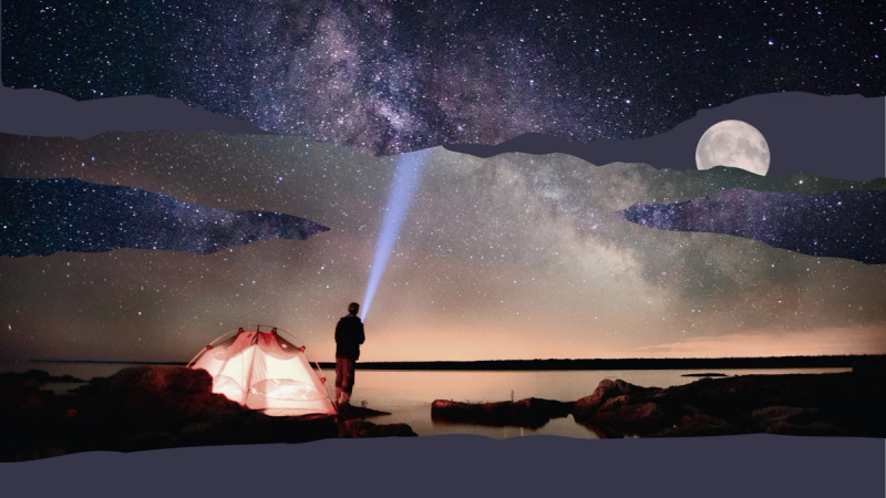A man standing by a lake pointing a flashlight into the starry night sky.