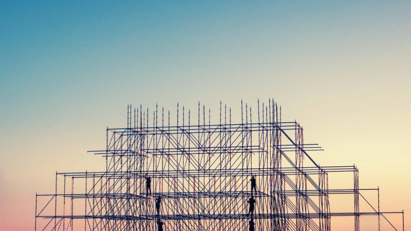 Tall scaffolding structure with workers in front of a orange to blue gradient sky.